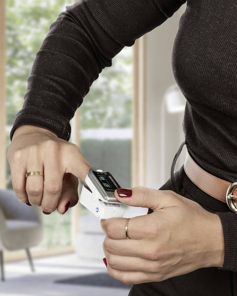 A person is holding a device to measure blood sugar. A bright living room is visible in the background.