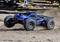 A blue remote-controlled monster truck speeds across a dusty surface, kicking up clouds of dust and travelling at high velocity.