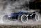 A remote-controlled car with blue and black colouring drives through dust on an off-road track. The vehicle has large tyres.