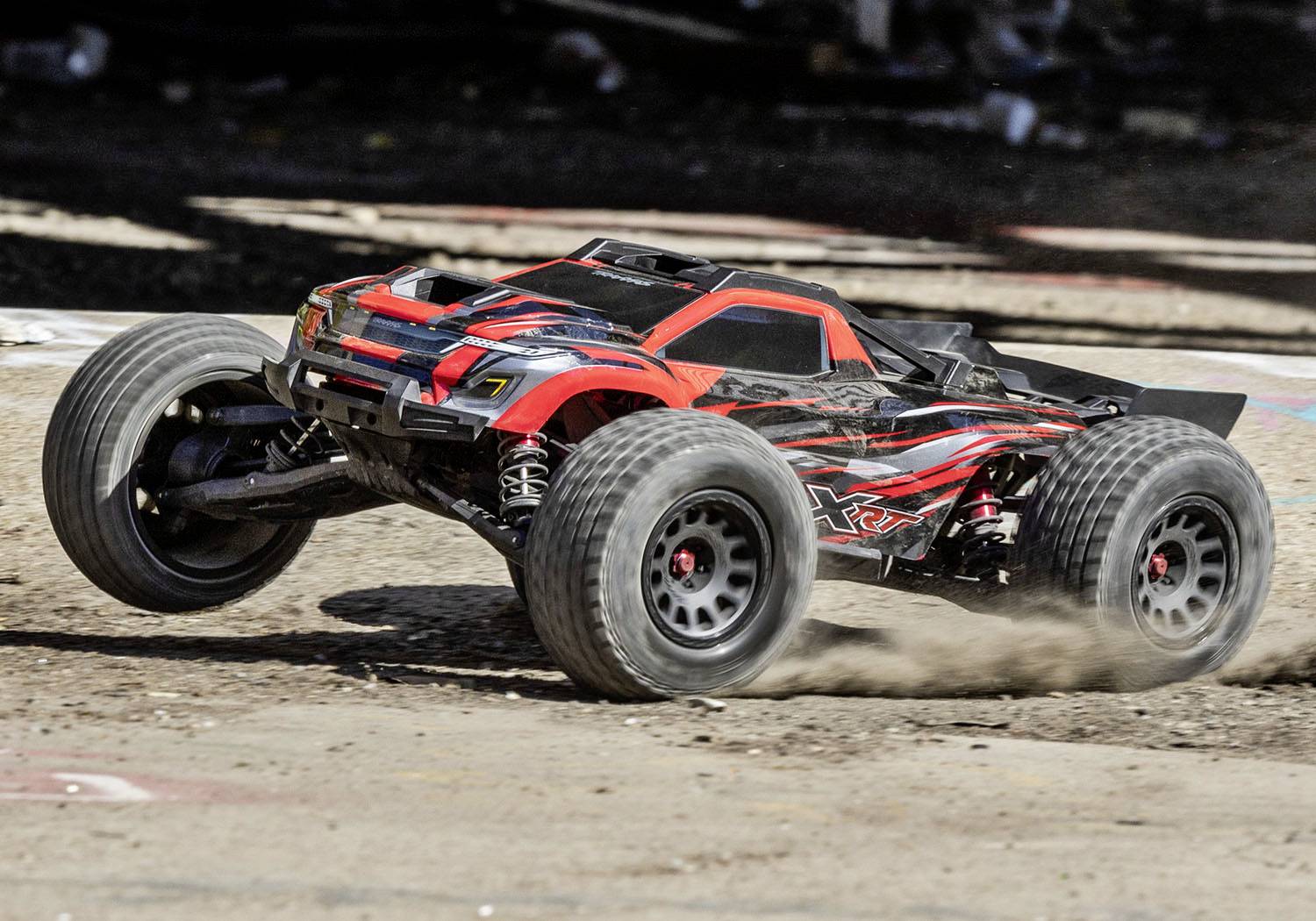 A red remote-controlled car is driving at high speed across an uneven, dusty track. Chunky tyres kick up dust.