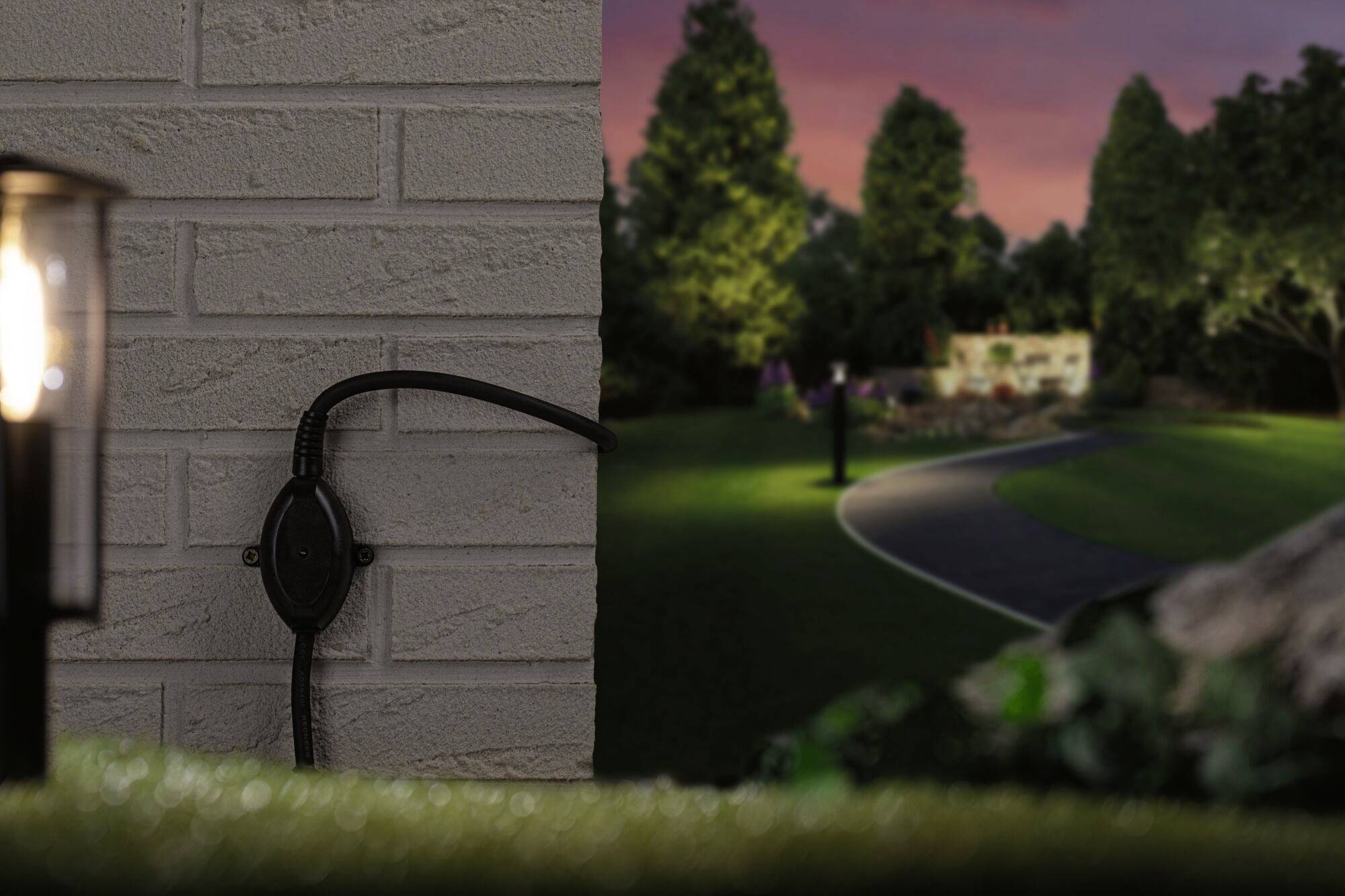 A illuminated garden at dusk; a lantern and a socket on a brick wall in the foreground; trees in the background.