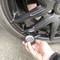 A hand is checking the tyre pressure of a car tyre with a tyre pressure gauge. The tyre is black and on asphalt.