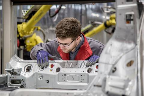 Man in workwear inspects automotive components in a factory, with a yellow robotic arm working in the background.
