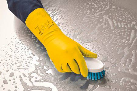 A hand wearing a yellow rubber glove is using a brush to clean a wet surface with soapy water.
