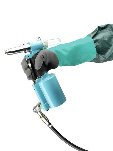 A hand wearing a green glove is holding a blue rivet gun. The device is connected to a compressed air hose.