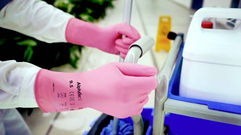 A person wearing pink gloves cleans a floor with a cleaning trolley, with plants and a warning sign visible in the background.