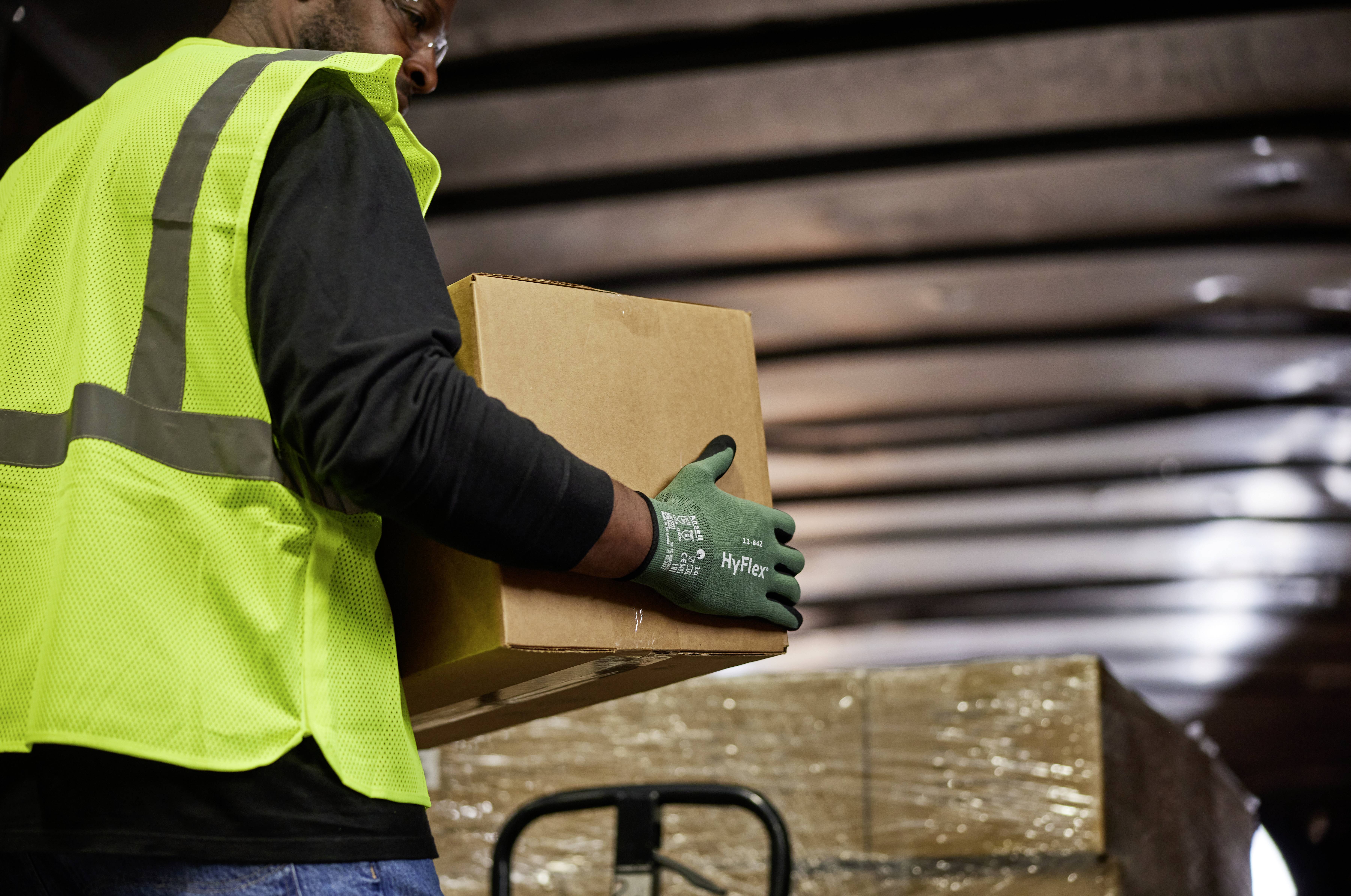 A person wearing a yellow high-visibility vest is lifting a cardboard box in a storage warehouse. Packaged pallets are visible in the background.