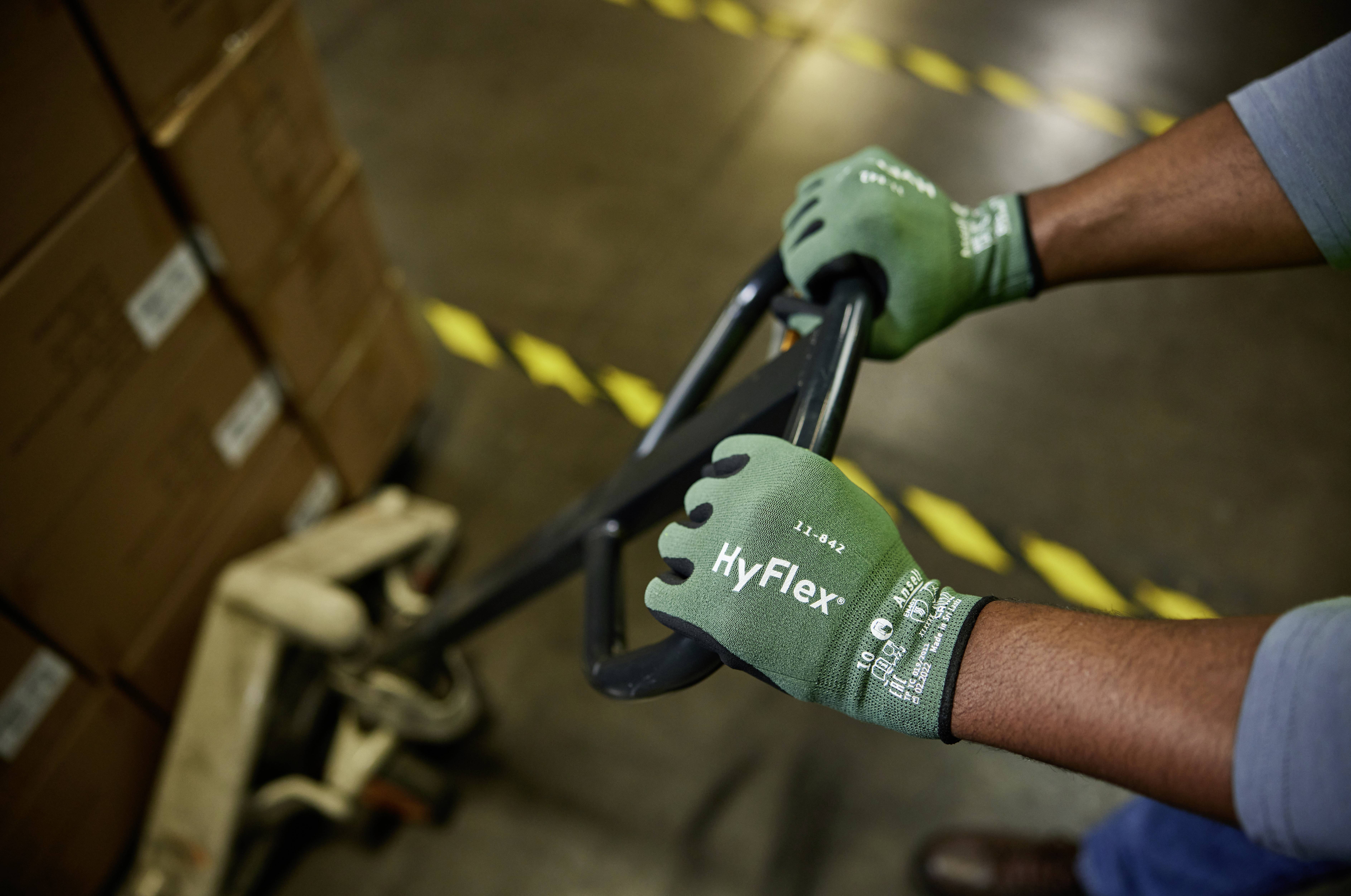 A person wearing green gloves operates a pallet truck in a warehouse to move boxes. Stacked cardboard boxes are visible in the background.