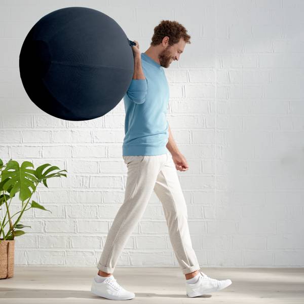 A man in casual clothing balances a black exercise ball on his back. A plant is visible in the background.