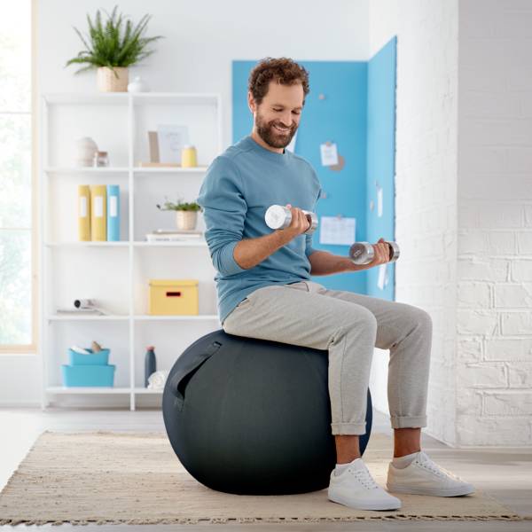 A man is training in the living room with dumbbells, sitting on a fitness ball, wearing comfortable sportswear. In the background, a shelf with plants.