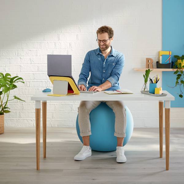 A person is sitting on an exercise ball at a desk, working on a laptop, surrounded by plants and colourful books.