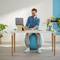 A person is sitting on an exercise ball at a desk, working on a laptop, surrounded by plants and colourful books.