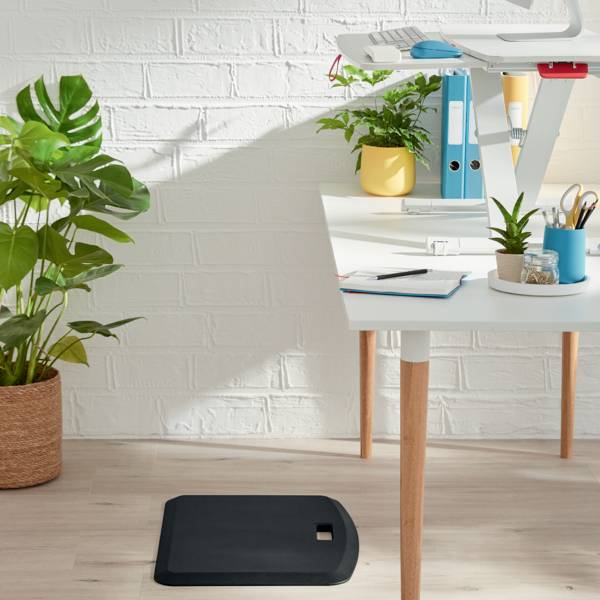 Desk with standing workstation, potted plants, folders and writing materials on light wooden flooring against a white wall.