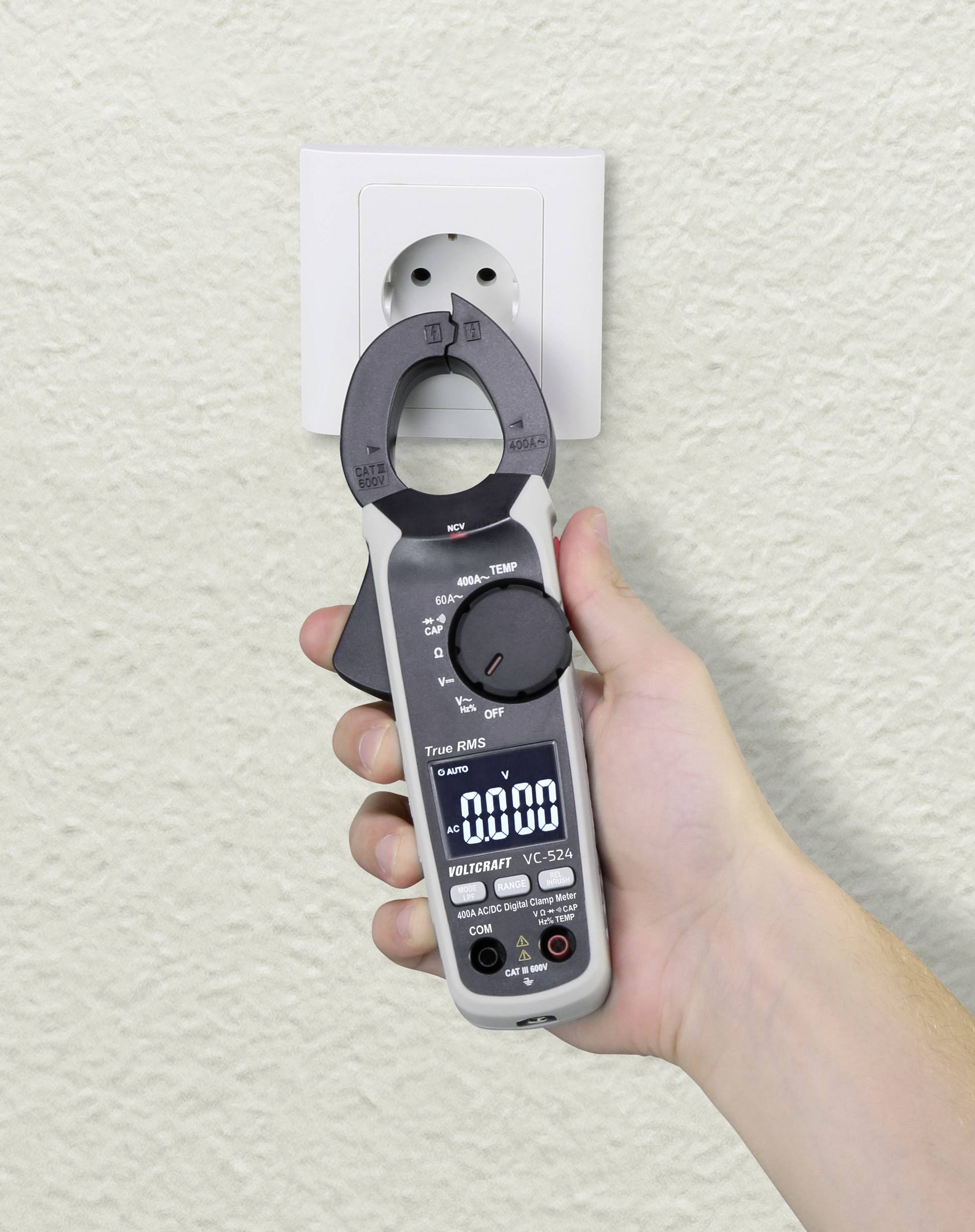 A hand is holding a digital electricity metre next to a wall socket. The display shows measurement readings, and the clamp is open.
