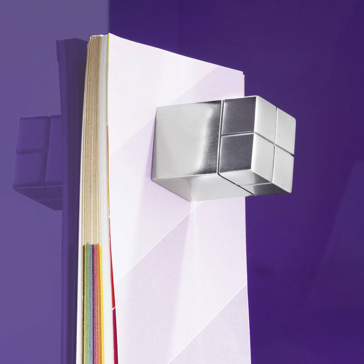 A cube magnet holds coloured, stacked paper sheets on a purple wall.