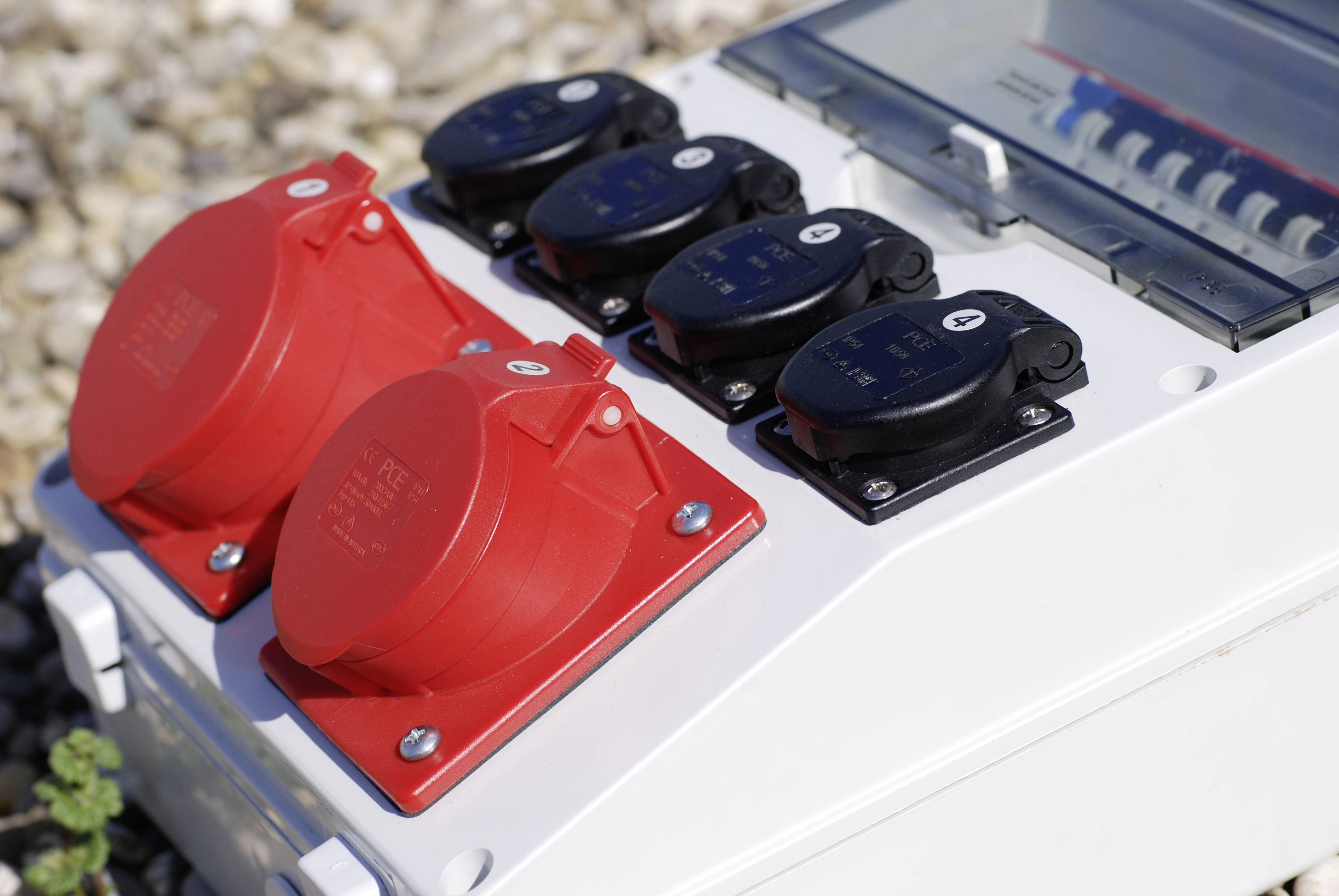 Several red and black power sockets on a white outdoor electrical distribution box, installed on a gravel surface.