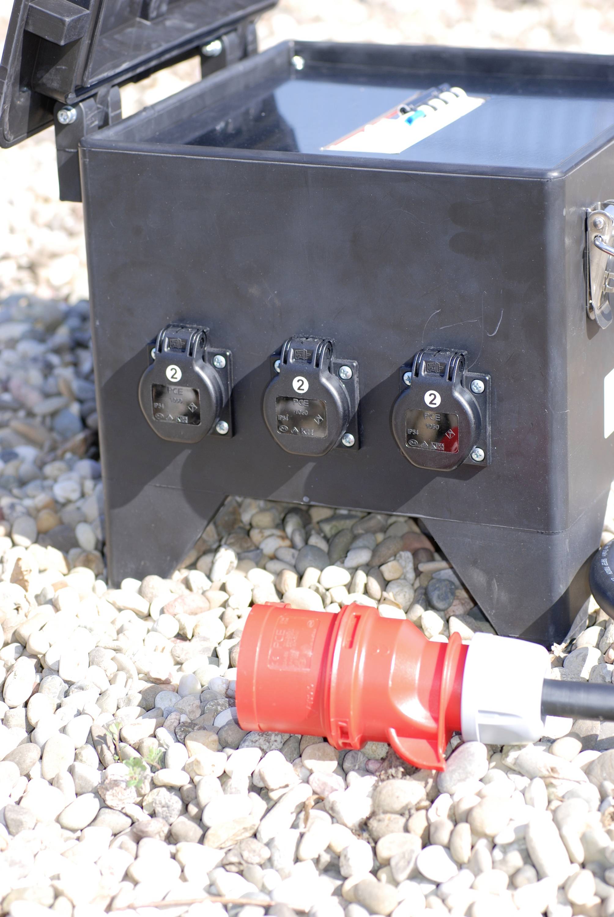 A black electricity distribution box with three sockets stands on gravel ground. A red plug lies in front of it.