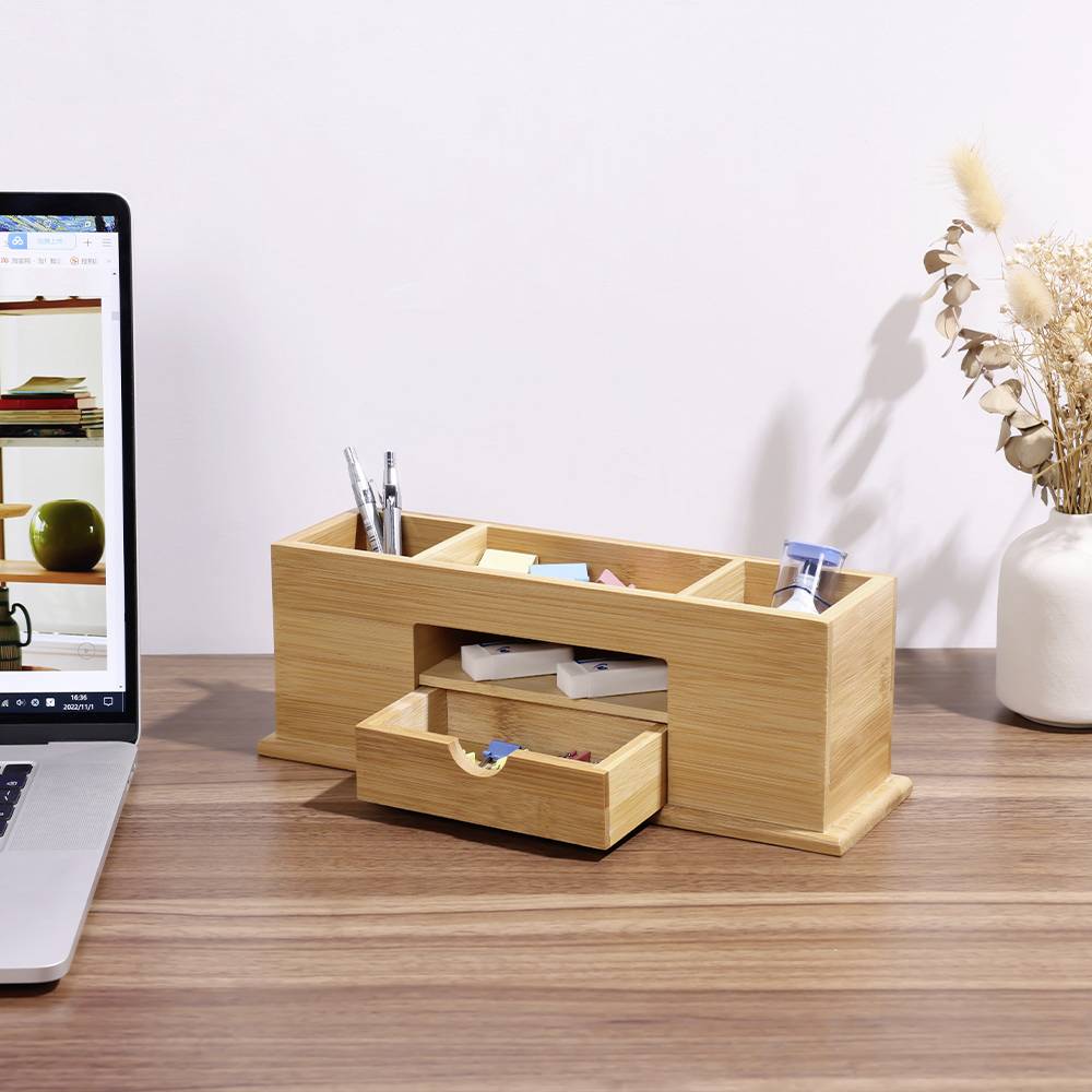 A wooden desk organiser with compartments and a drawer sits on a table next to a laptop. A vase with dried flowers is placed beside it.