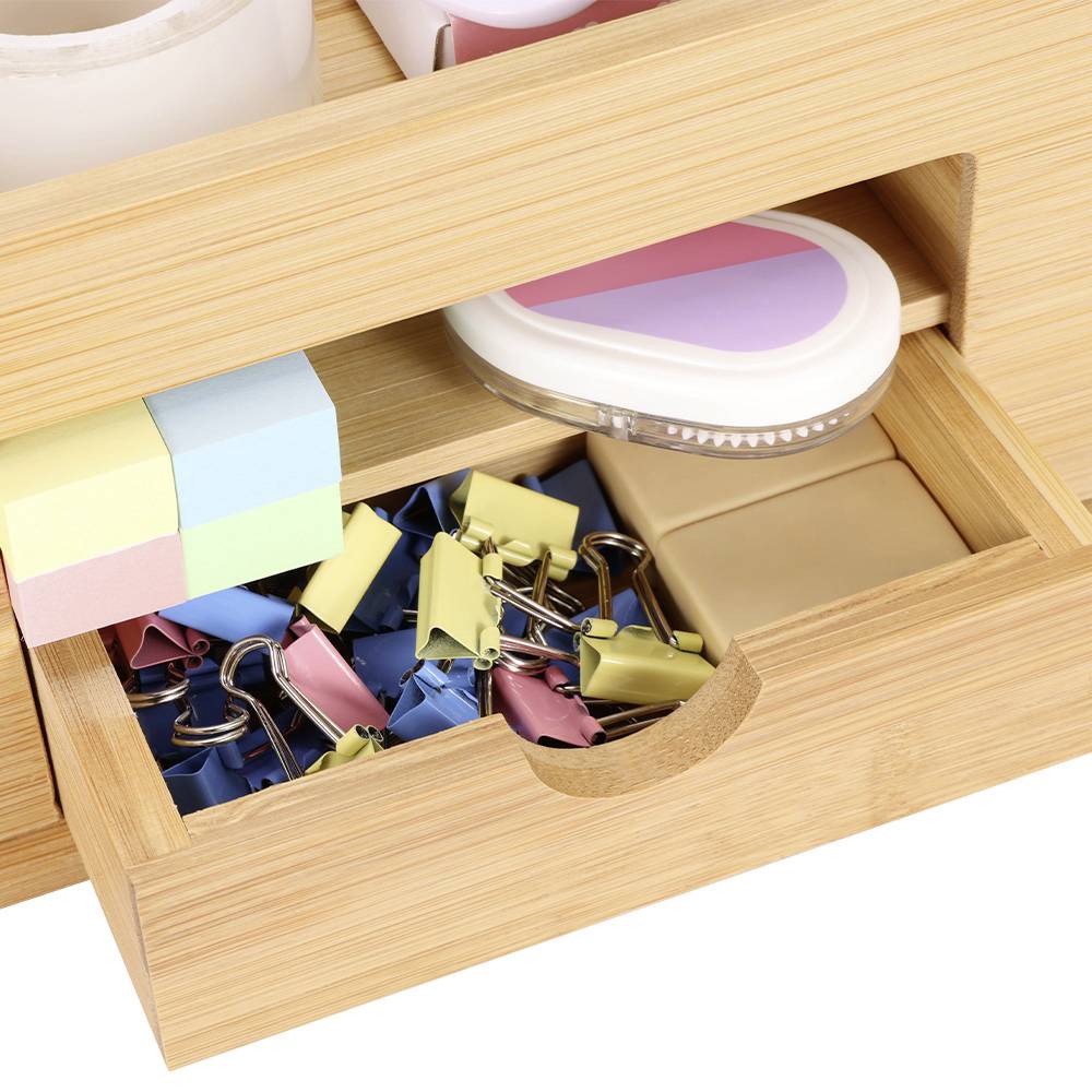 An open wooden drawer filled with colourful paperclips, notepads, and office supplies. A folder is lying on the shelf above.