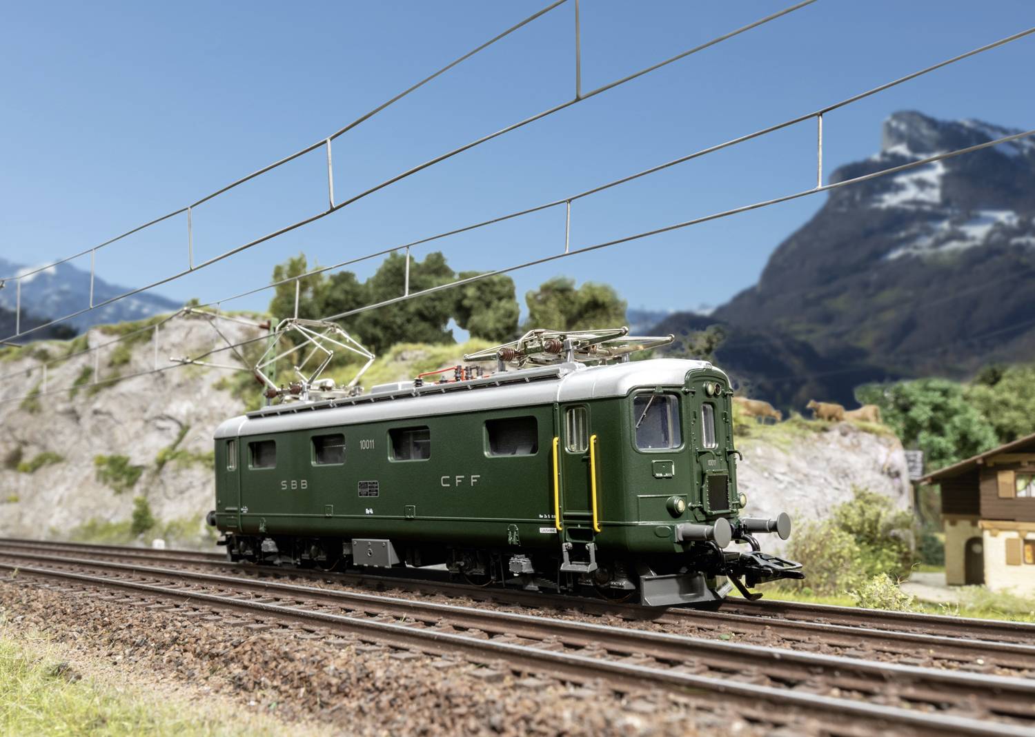 A green train travels on tracks through a mountainous landscape. Power lines run overhead. Mountains are visible in the background.