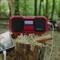 Red portable radio with display showing FM station '103.0'. Sits on tree stump in forest. A smartphone with headphones lies beside it.