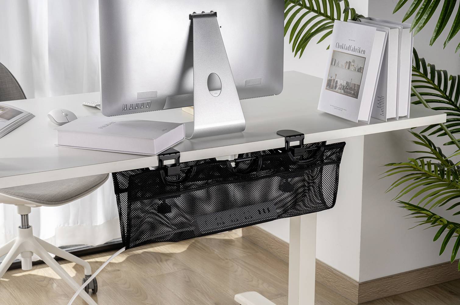 A desk with a computer and cable management basket underneath. Books are lying on the table, and plants can be seen in the background.