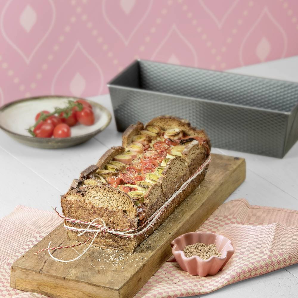 A freshly baked loaf with tomatoes and leeks on a wooden board, alongside a bowl of cherry tomatoes and a bread tin.