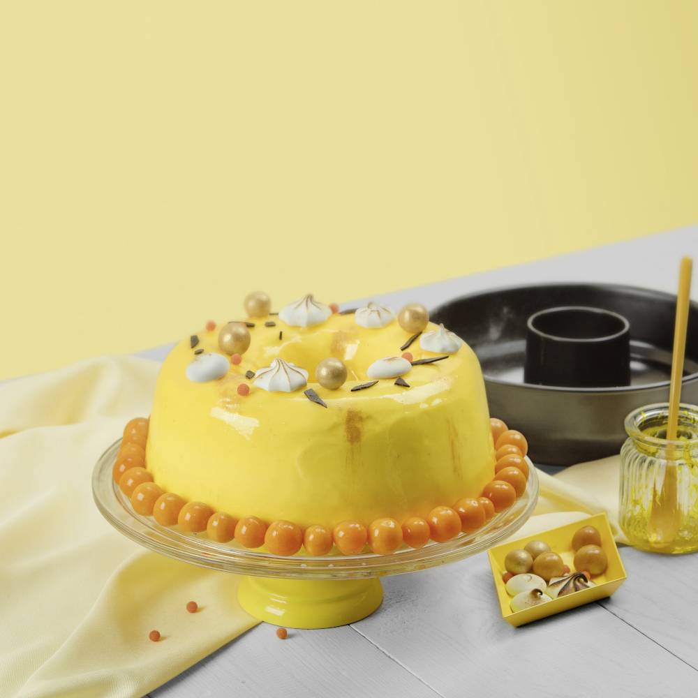 A yellow cake with a glossy coating, decorated with golden spheres and white foam, sits on a yellow cake stand.