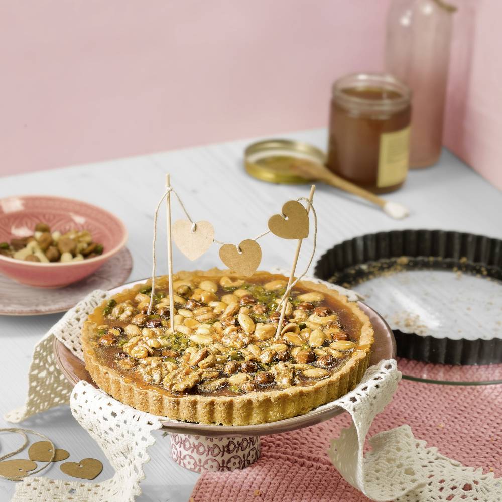 A tart decorated with nuts and seeds sits on a table, surrounded by empty plates and a glass of honey in the background.