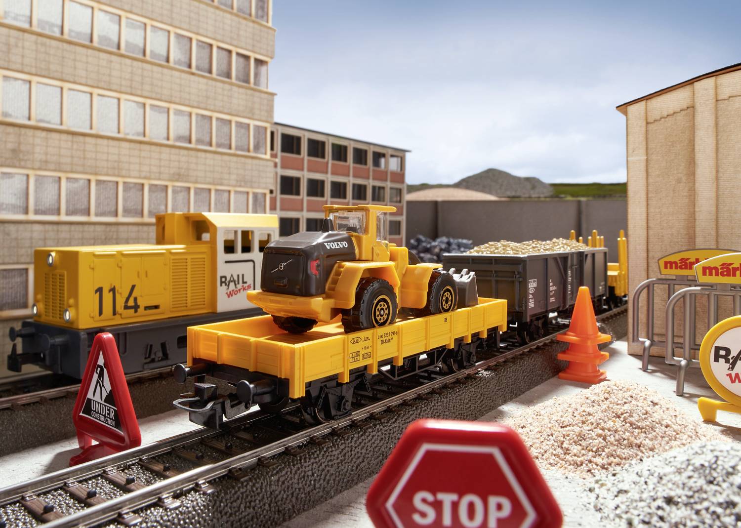 A model train with a yellow locomotive and goods wagons is transporting a toy construction machine. Buildings can be seen in the background.