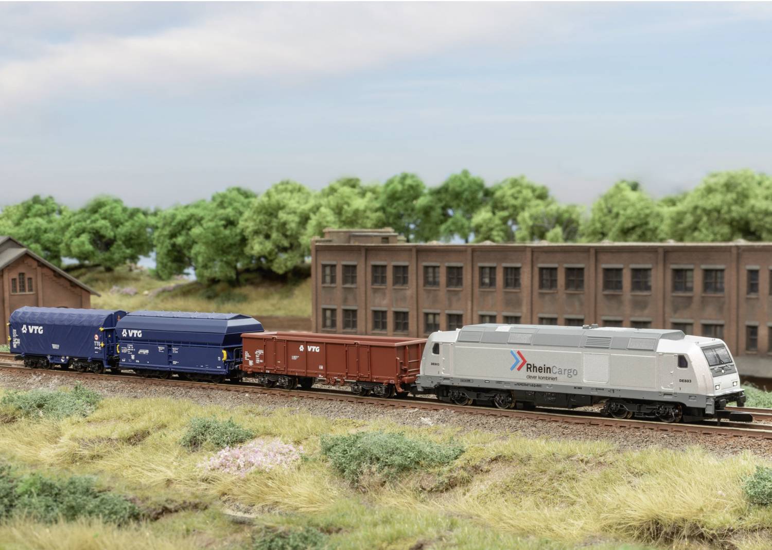 A model train with a grey locomotive and two blue carriages travels alongside a brick building in a rural setting.