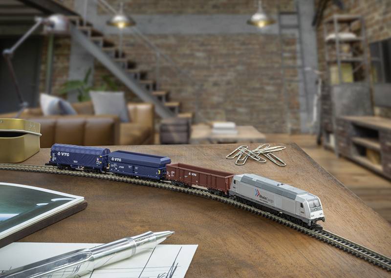 A small model train with several cars is set up on a wooden table in an industrial-style living room with a staircase and leather sofa.