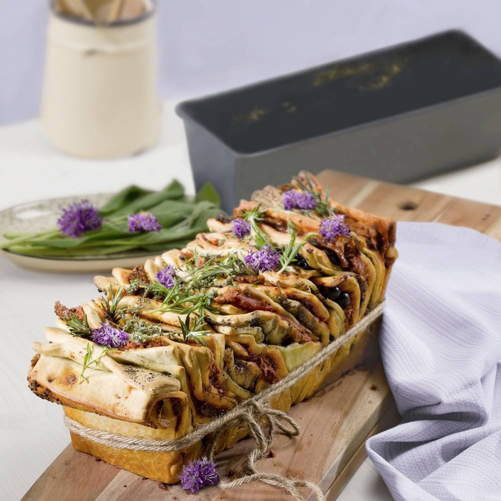Freshly baked bread with herbs and purple blooms on a wooden board, alongside a black baking dish and a plate.