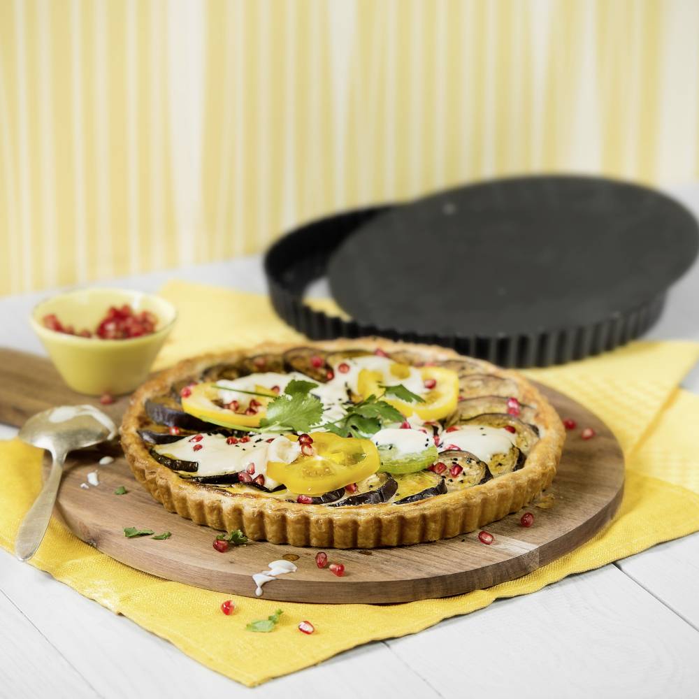 A vegetable quiche with courgettes, tomatoes and pomegranate seeds, garnished with herbs, on a wooden board against a yellow background.