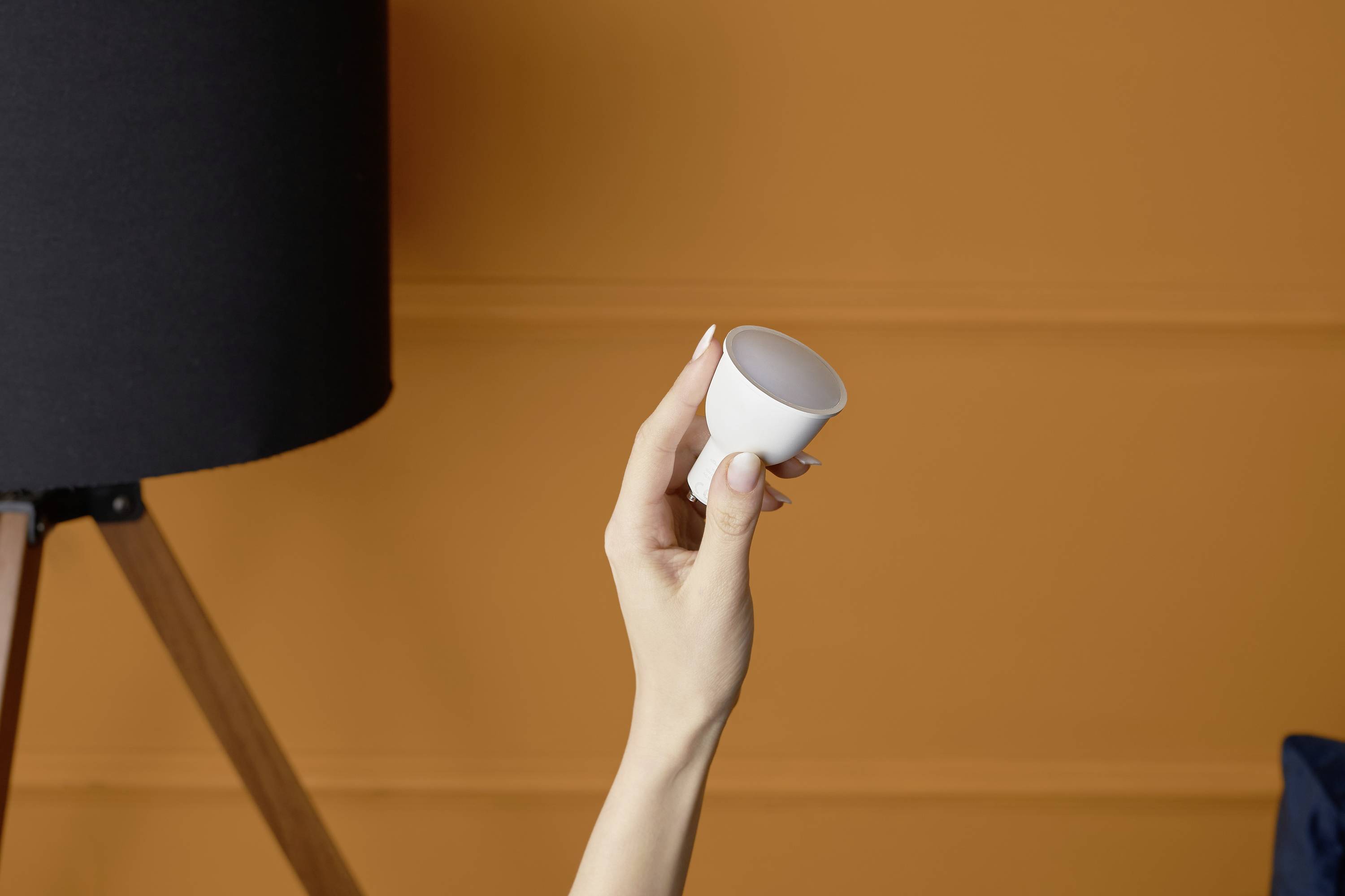 A hand holds a small white cup in front of an orange background. A black lamp stands on wooden legs to the left.