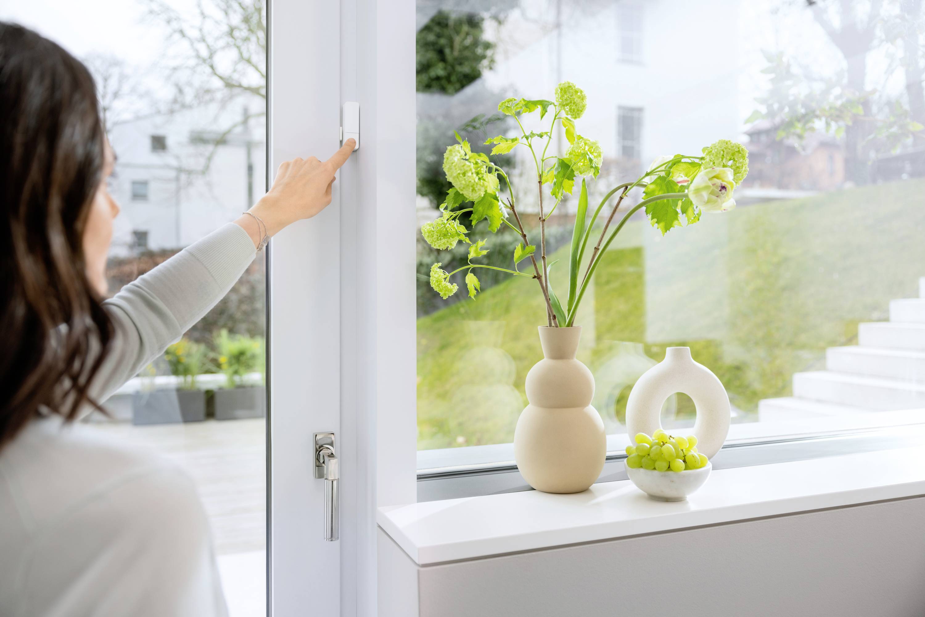 A woman opens a window. On the windowsill are a vase of flowers and a bowl of green grapes.