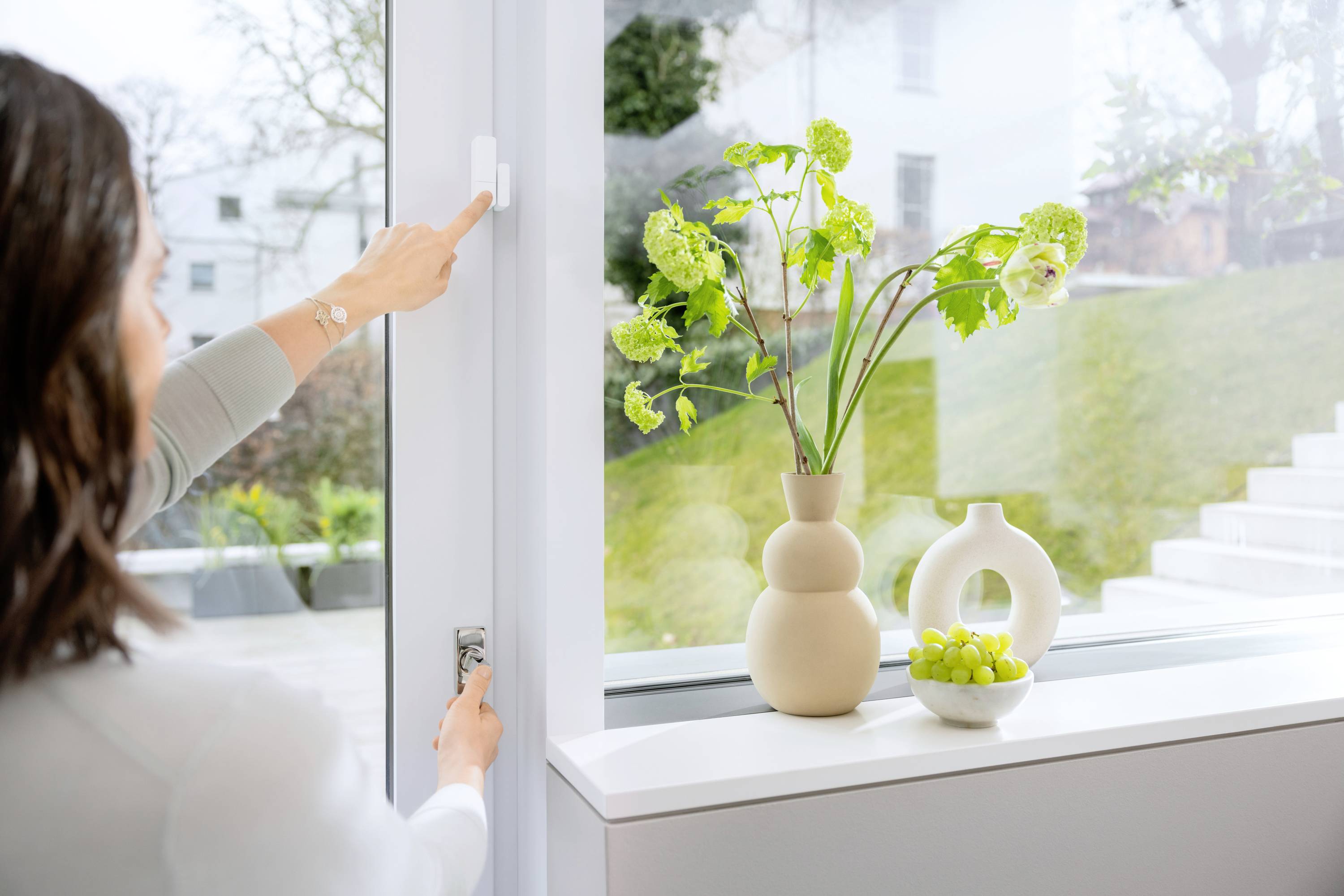A person opens a window; on the windowsill stands a vase with flowers and a bowl of green fruits.