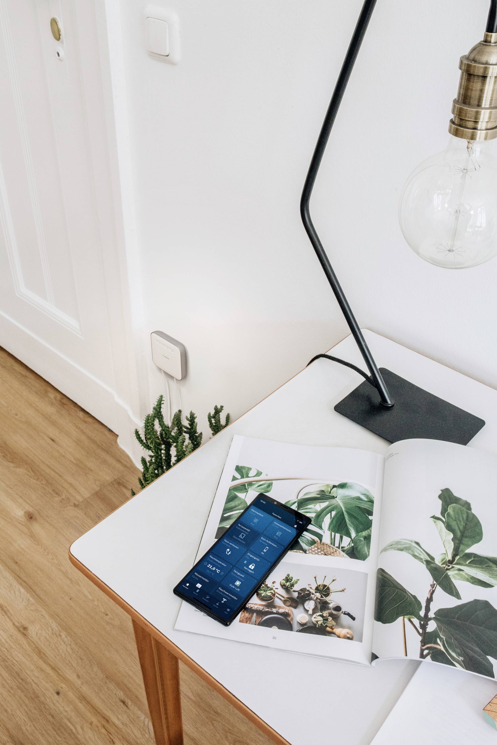 A smartphone lies on a table beside an open magazine with plant images and a lamp.