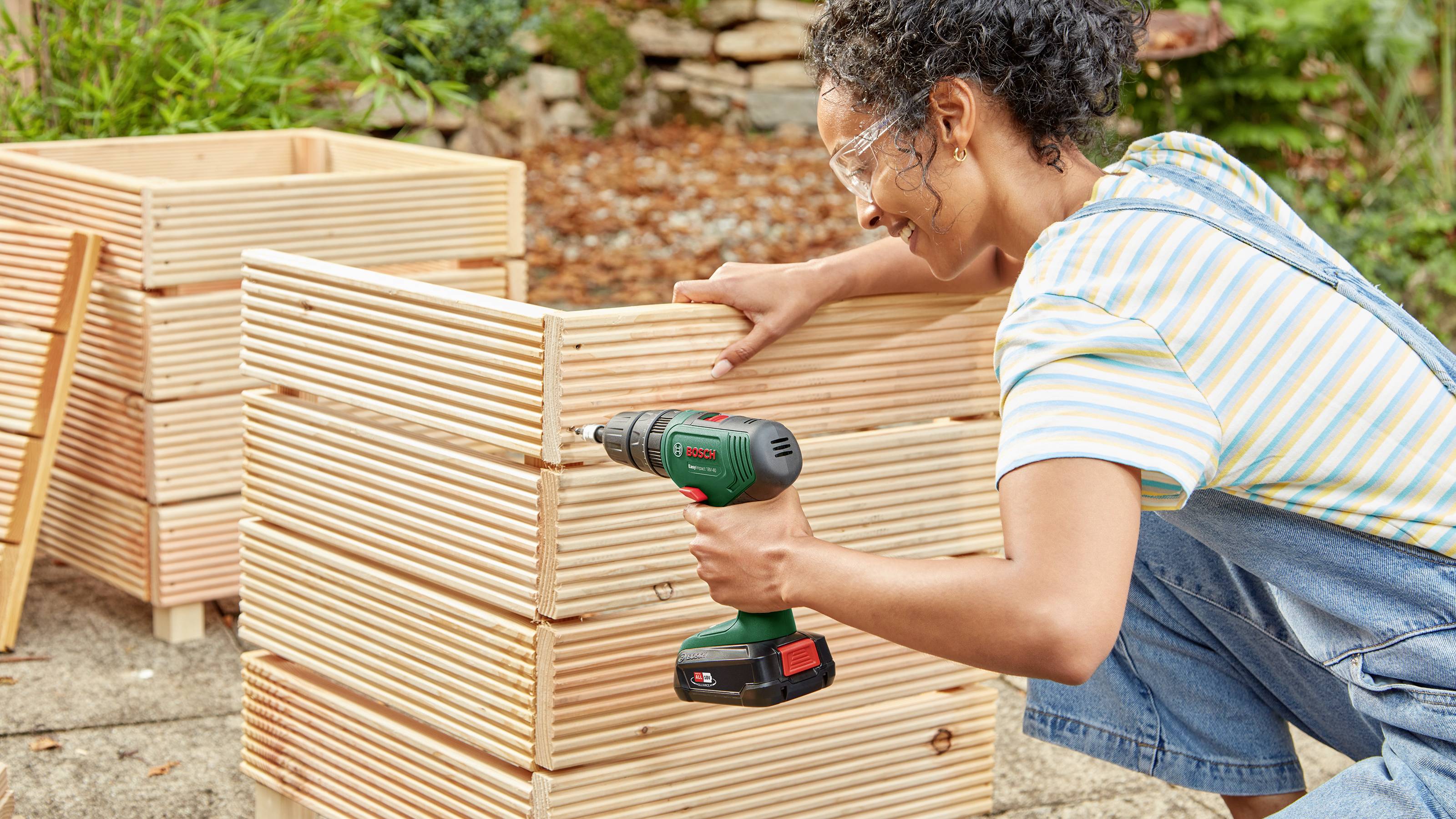 A person is constructing a garden bed made of wooden slats using a green drill. Further wooden crates are visible in the background.