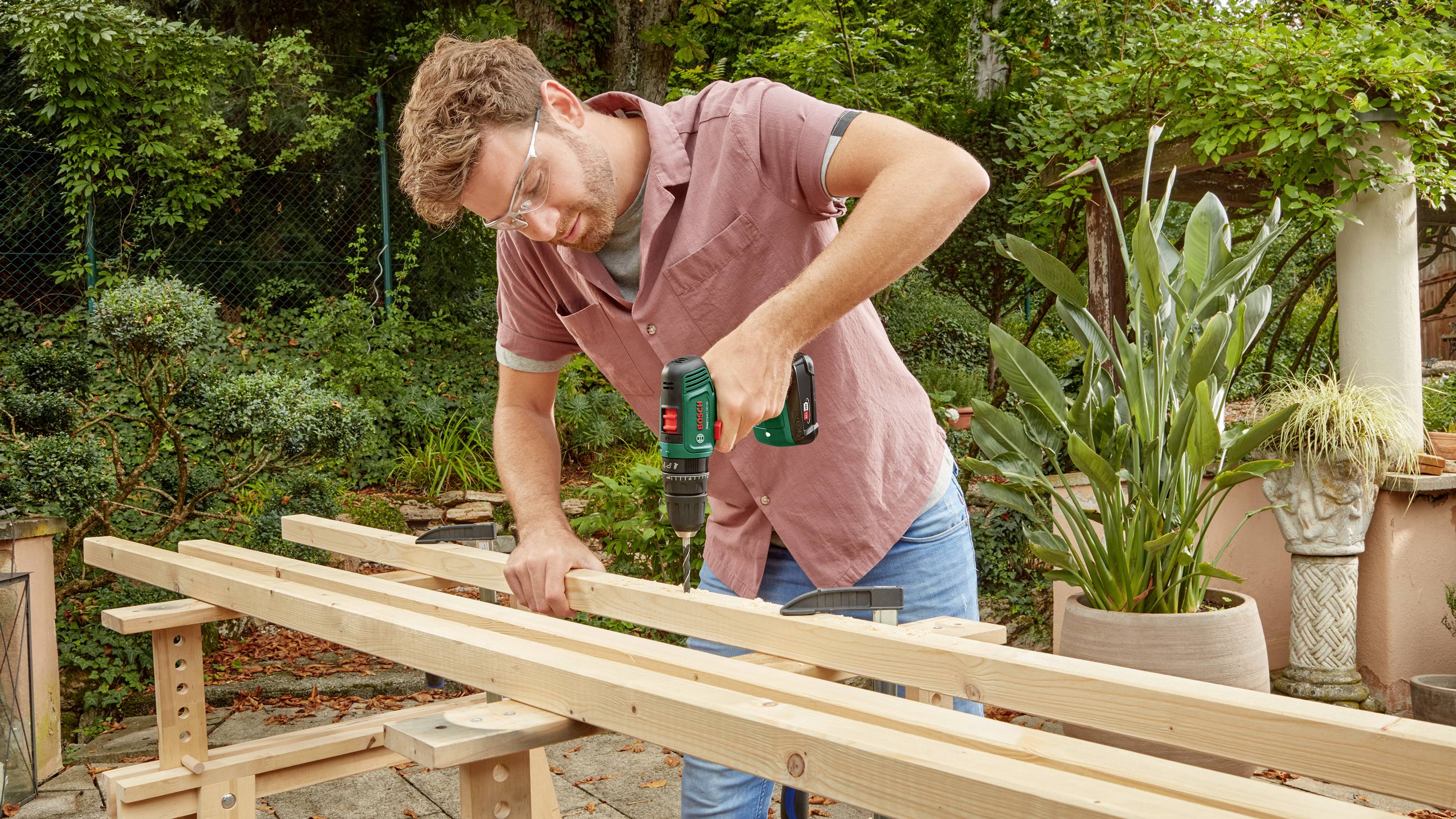 A man in a garden is drilling into a long wooden board with an electric drill, which is positioned on a workbench.