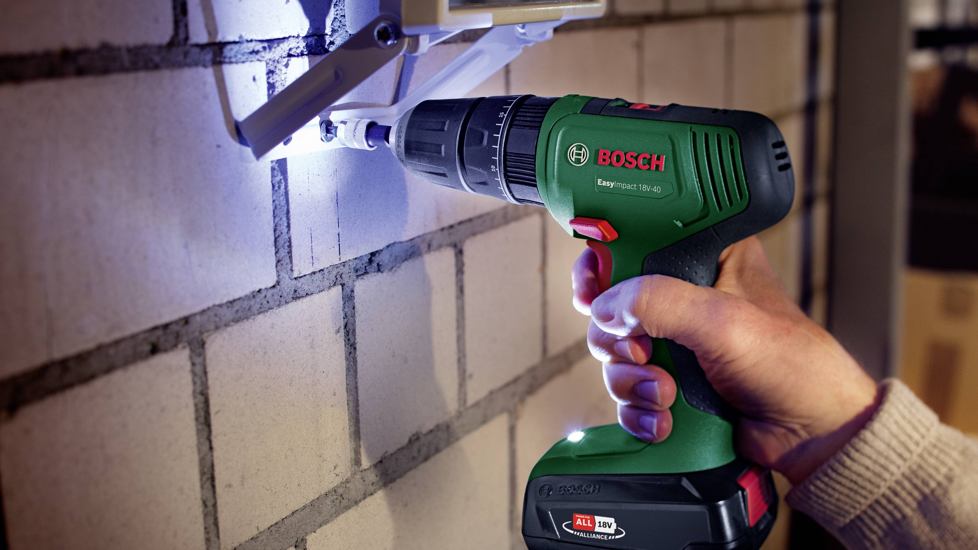 A person is using a green Bosch cordless drill to bore into a brick wall. Additional tools are visible in the background.