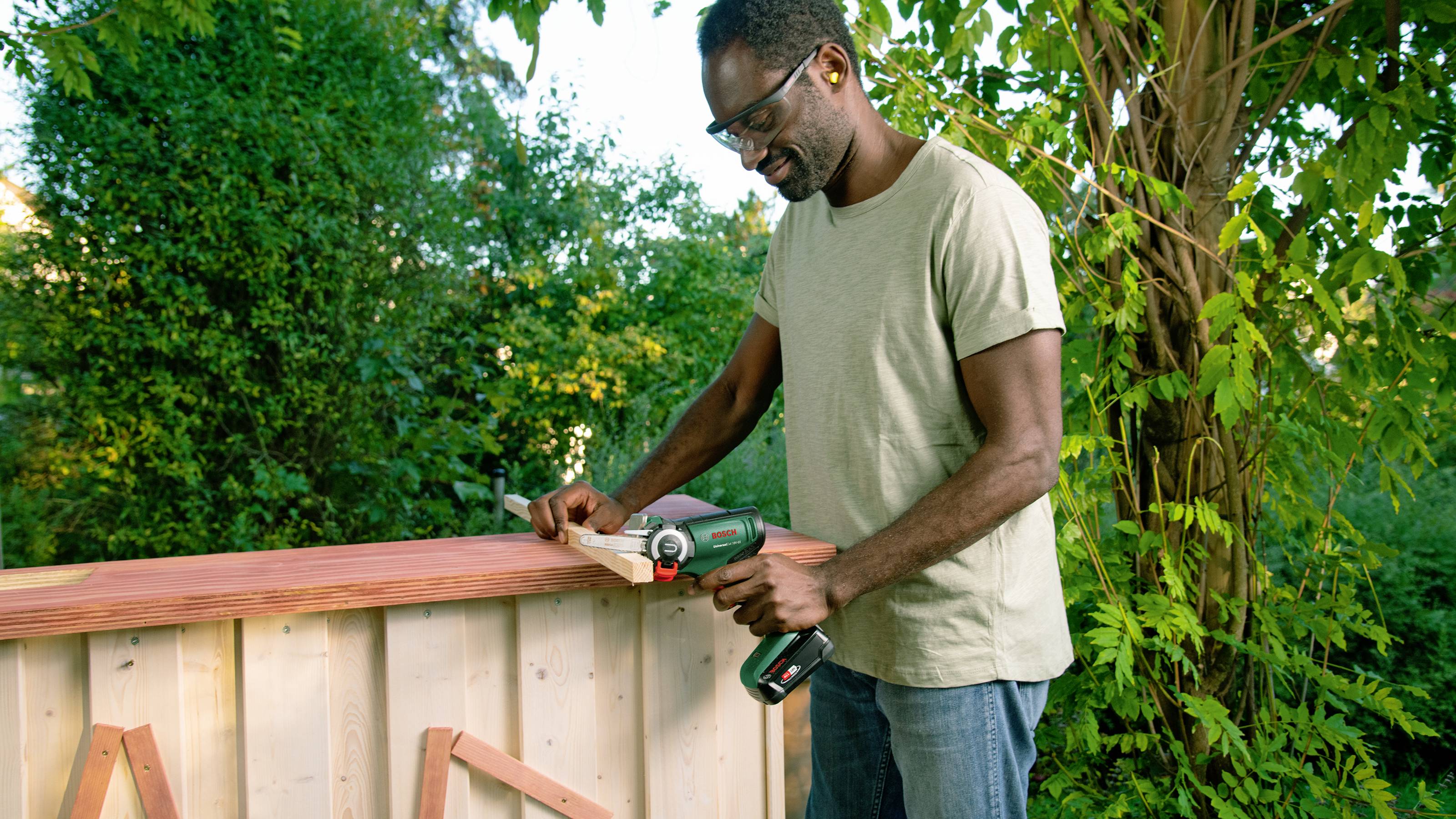 A man is working on a wooden project in a garden, using an electric drill to secure parts of a fence.
