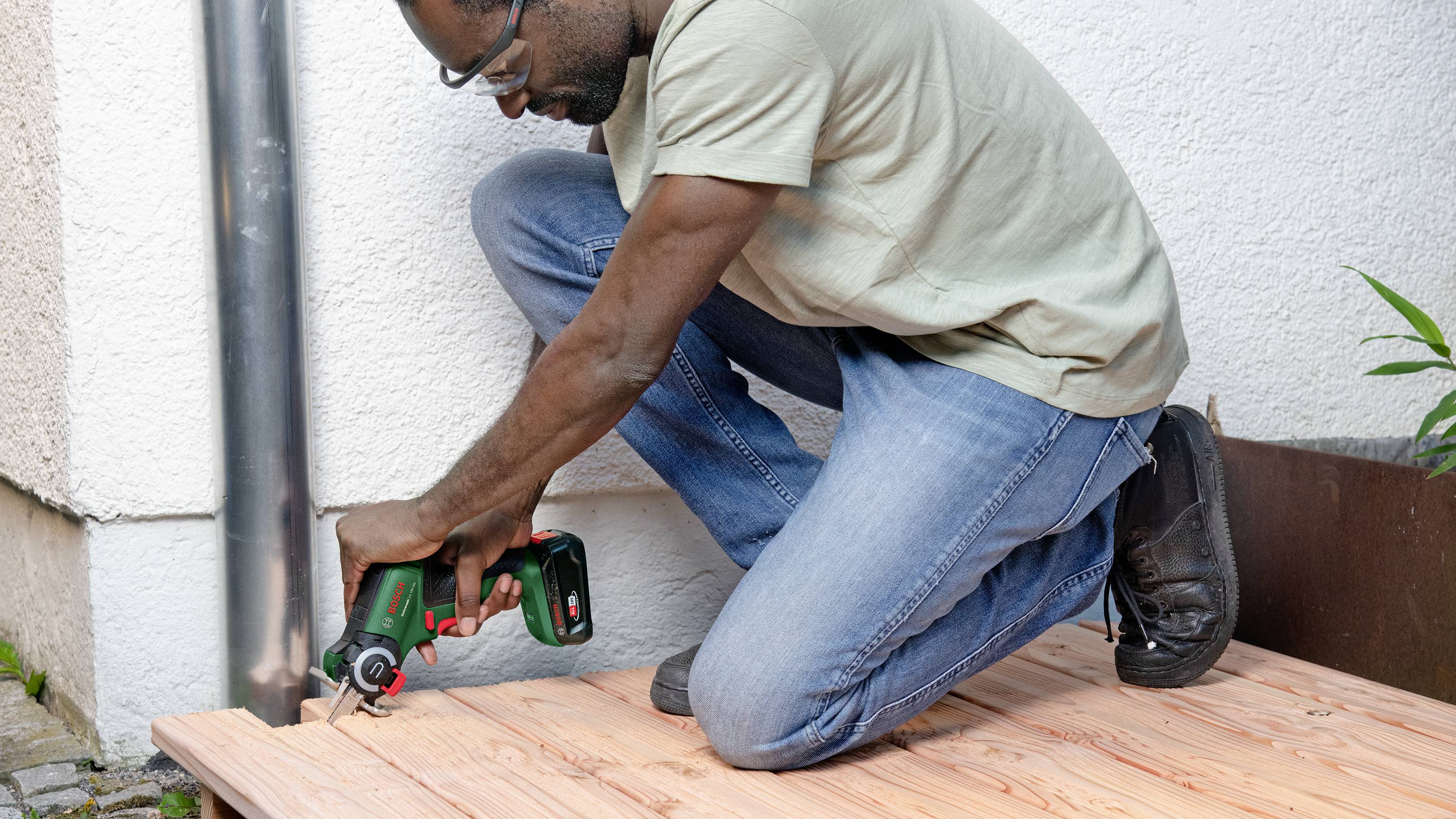 A person is kneeling on a wooden deck and using a green circular saw to cut the wood.