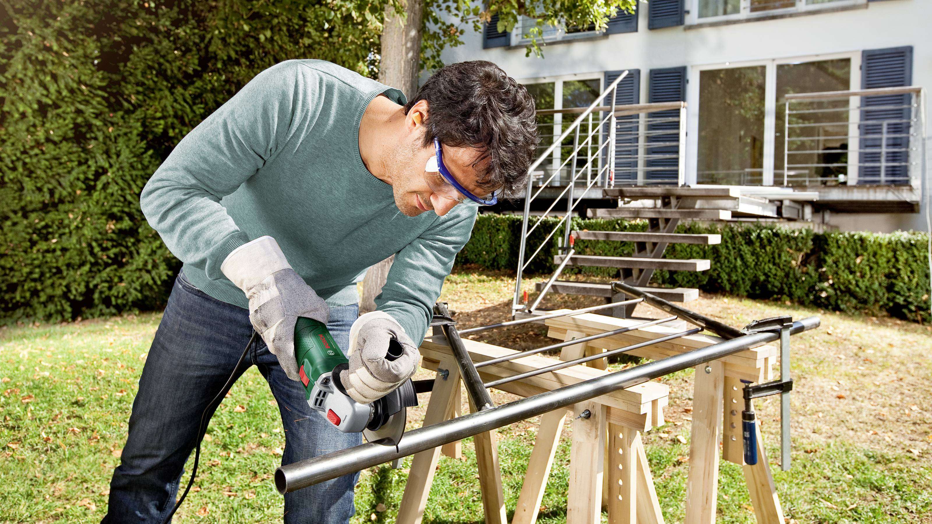 A man is cutting a metal pipe with an electric saw in the garden. A modern house can be seen in the background.