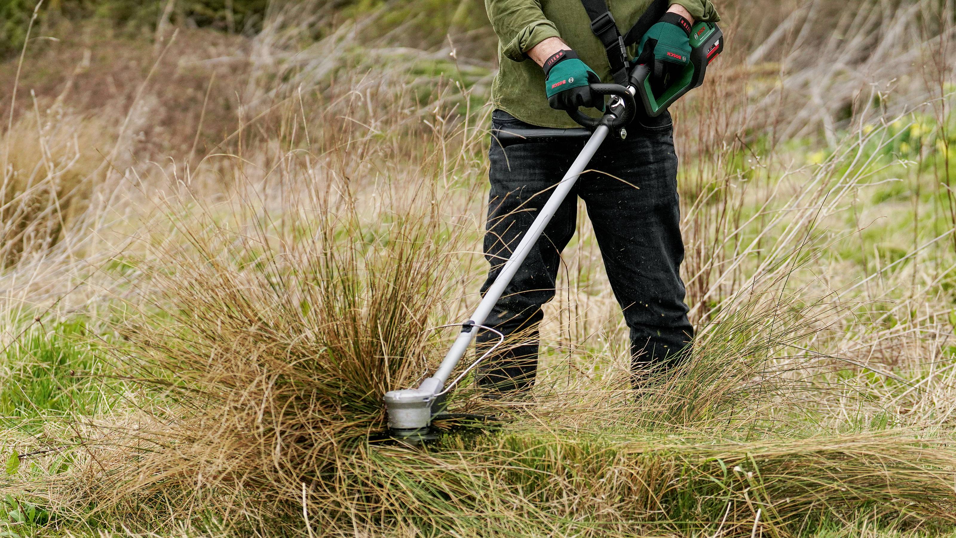 A person is cutting grass with a strimmer in an overgrown garden. Gloves protect the hands.
