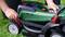 A person adjusts the cutting height of a green lawnmower. Hands are operating a red lever on the wheel on green grass.