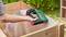 A person is stapling fabric to a wooden frame outdoors using a green electric staple gun.