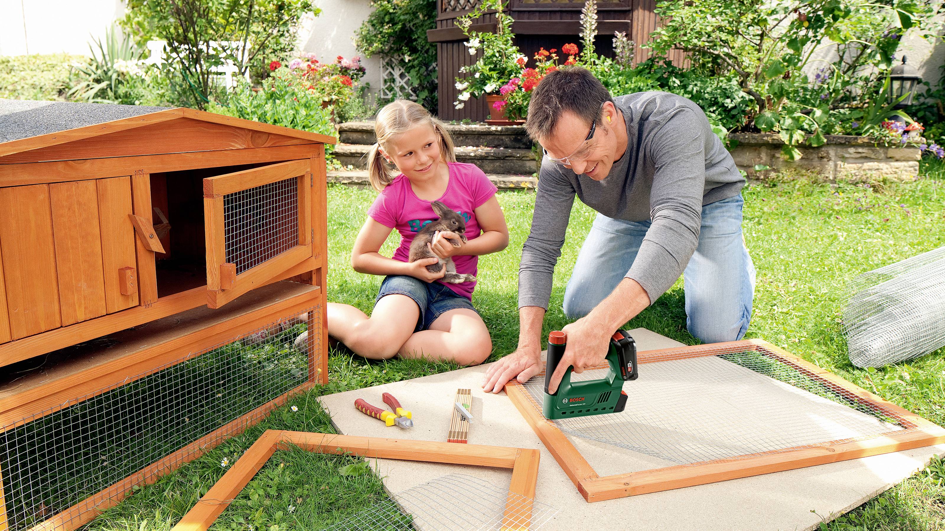 A man is building a wooden lattice next to a large rabbit hutch in the garden with a cordless drill. A girl is holding a rabbit.