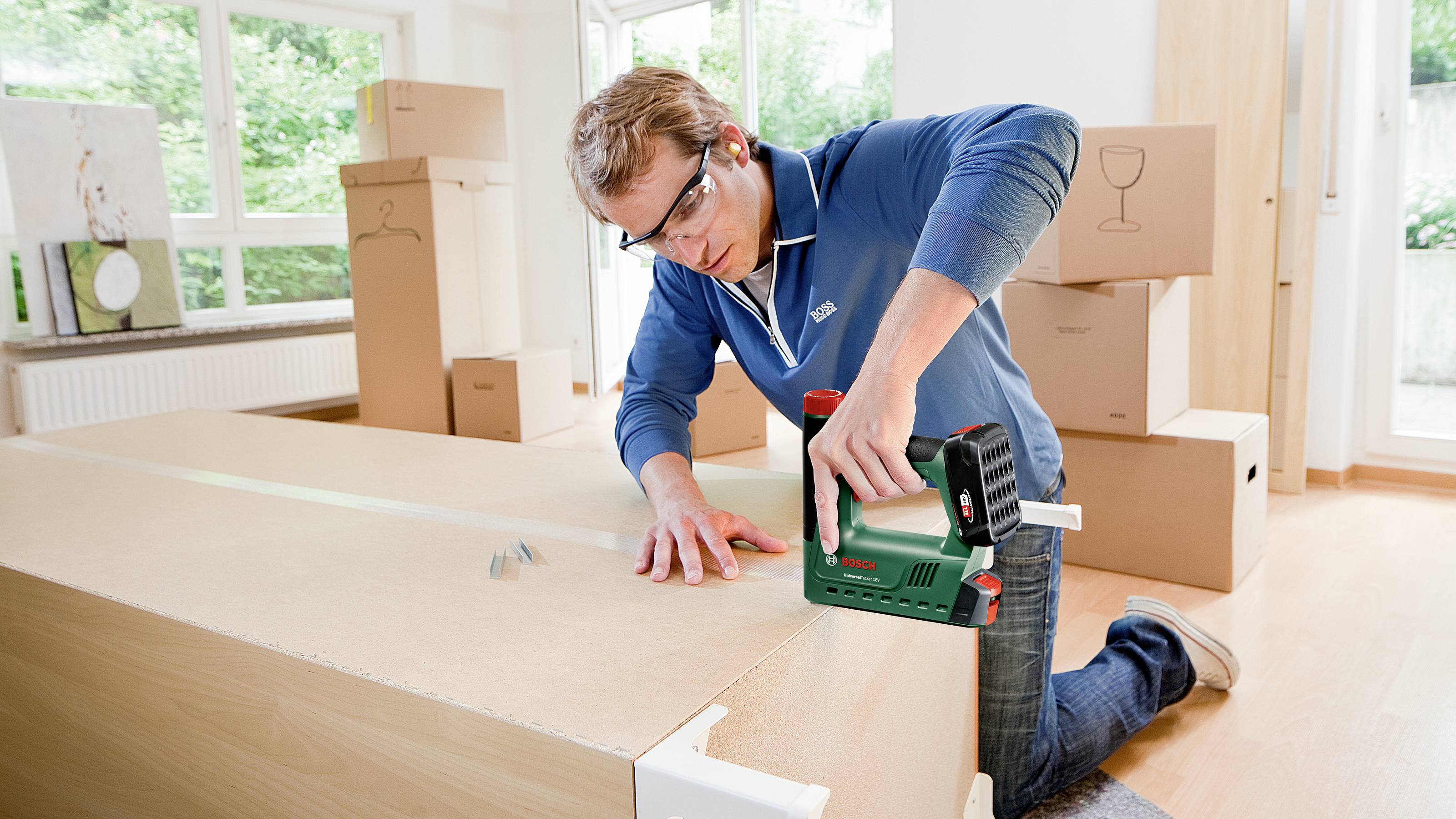 A man is assembling a piece of furniture. He is wearing safety glasses and using an electric power tool. Moving boxes can be seen in the background.