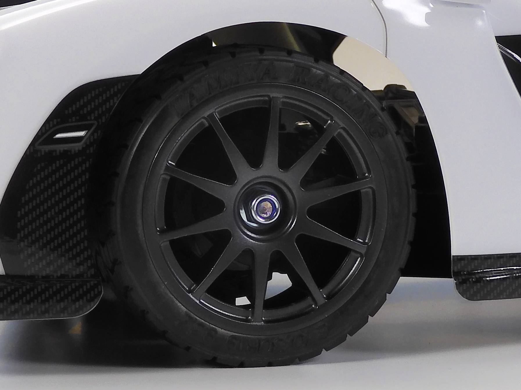 A section of a black car wheel with a multi-spoke rim on a white car, against a light background.
